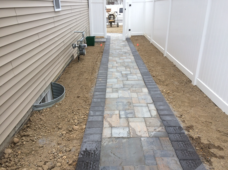 Brick walkway between a house and a white fence; unfinished landscaping.