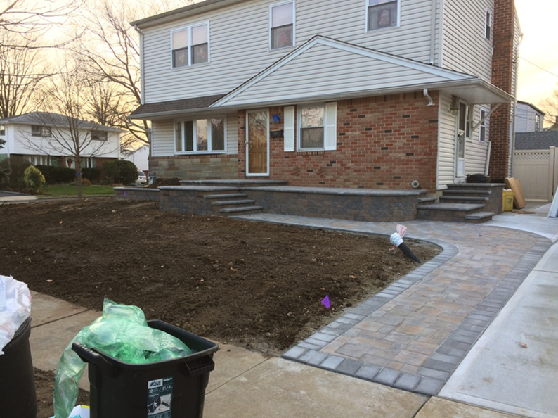 Two-story house with brick facade and paved walkway under construction, front yard with bare soil, trash bins visible.
