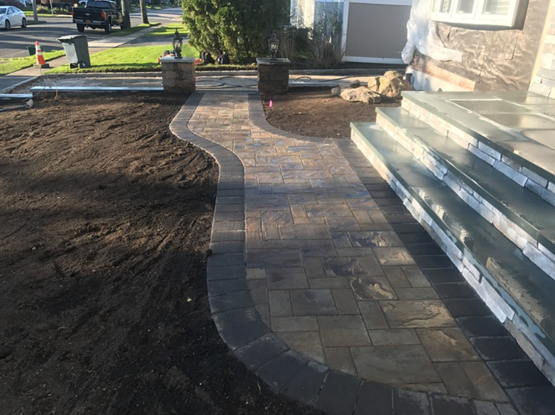 A paved walkway curves to a home's steps. Dark pavers with a black border. Soil and grass surround.