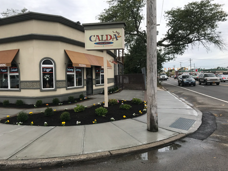 Exterior of Calda restaurant on a corner lot, featuring a sign, tan building, flowers, and a busy street.