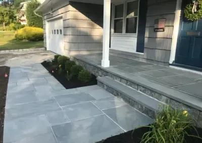 Gray stone walkway and porch, dark blue door, white column, garage, and plants.