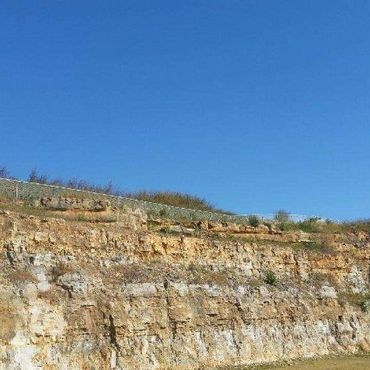 Layered rock cliff under a clear blue sky with sparse vegetation and a fence at the top.