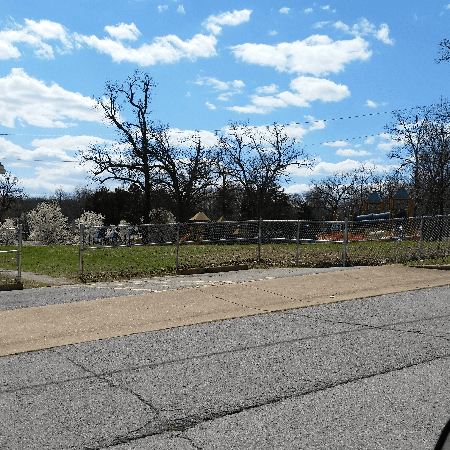 A sunny outdoor scene with a playground behind a chain-link fence, under a partly cloudy blue sky.