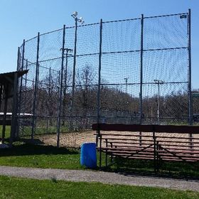 Baseball backstop with netting, bleachers, and a blue trash can. Field, trees, and sky in background.
