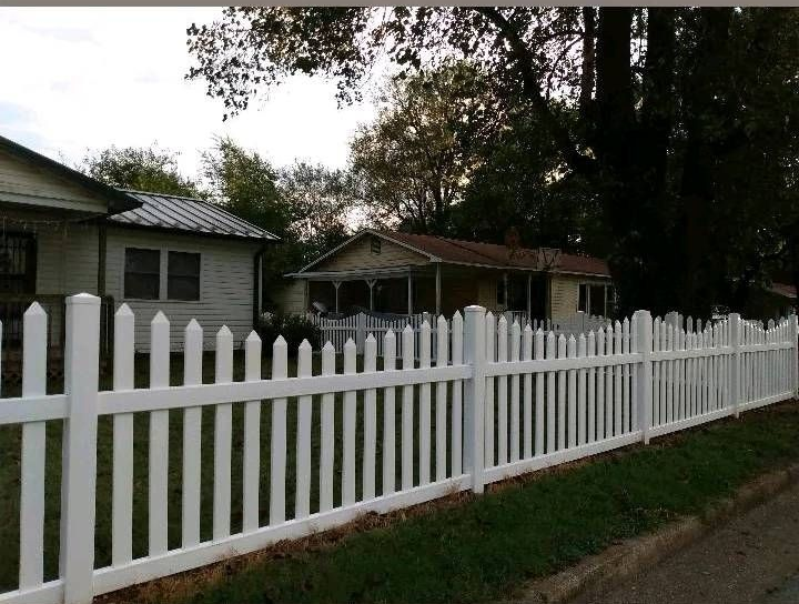 White picket fence along a grassy yard beside small houses and trees on a cloudy day