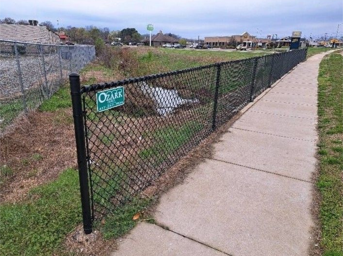 Chain-link fence along a sidewalk beside a grassy ditch under a cloudy sky