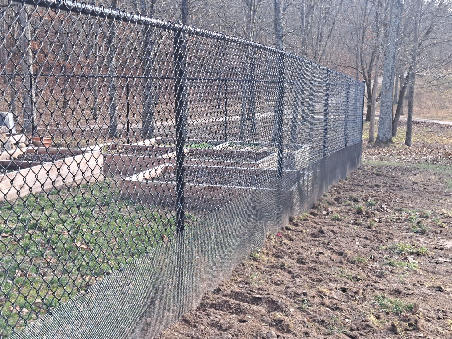 Chain-link fence beside a muddy, leafless wooded path with outdoor enclosures in the background
