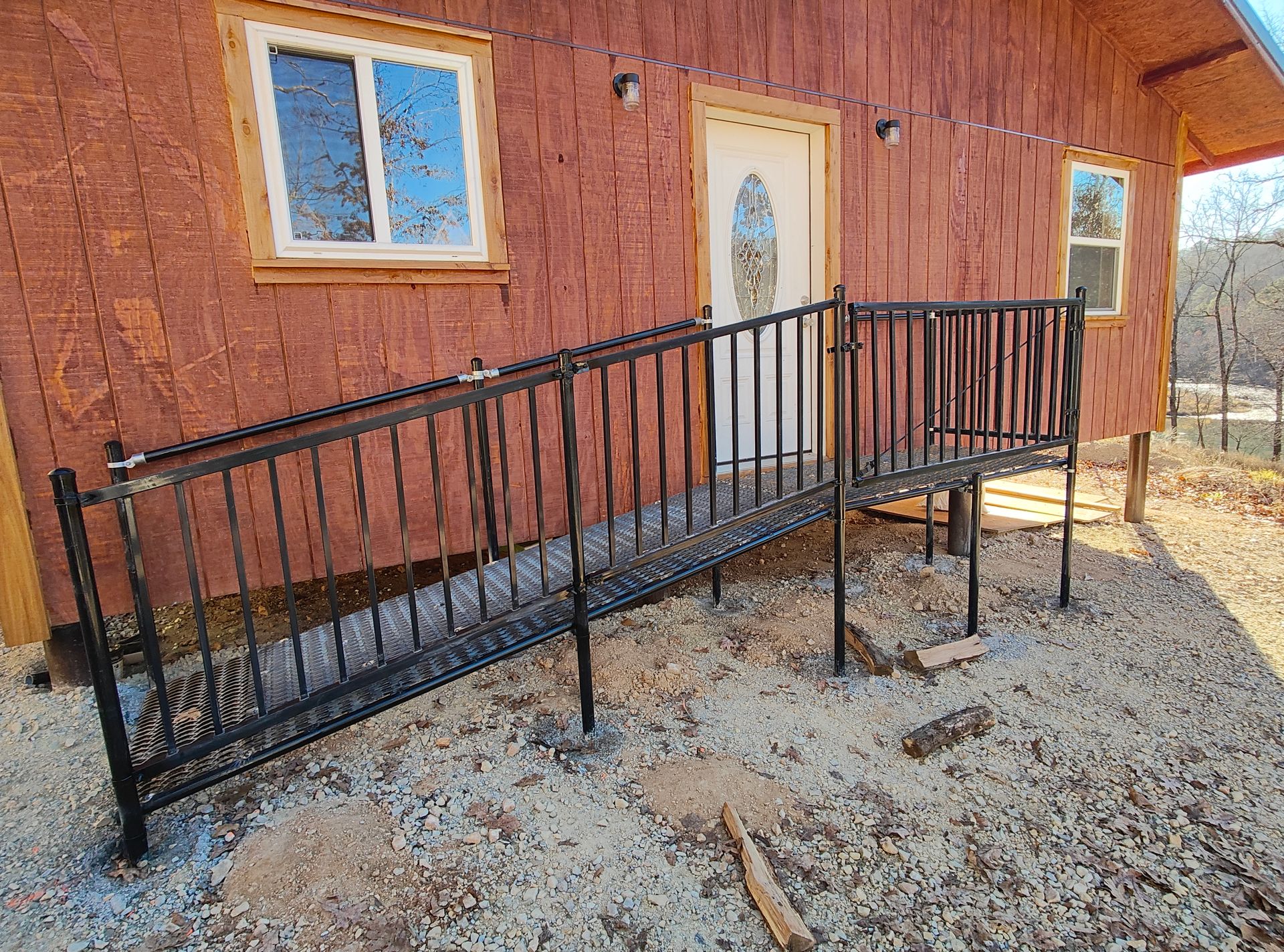 Red cabin porch ramp with black metal railings, white door, and gravel ground.
