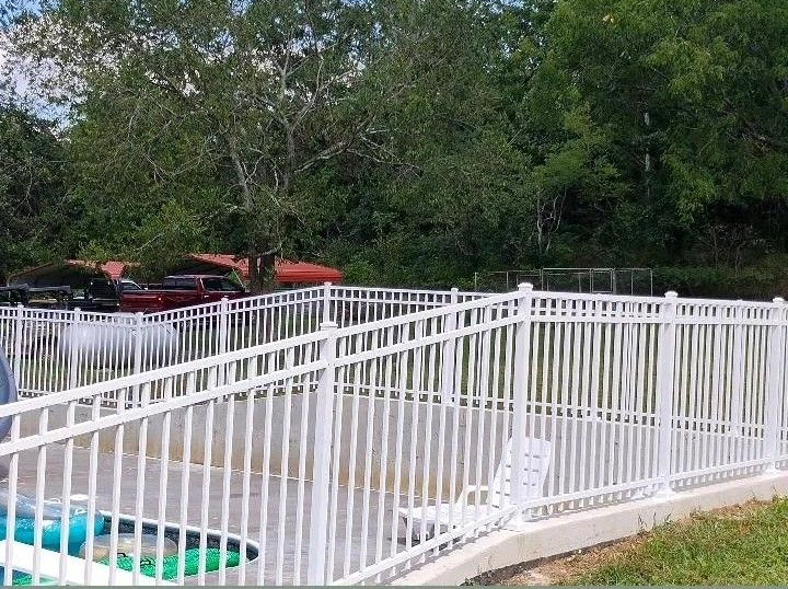 White fenced pool area with lounge chairs beside trees and parked cars in the background