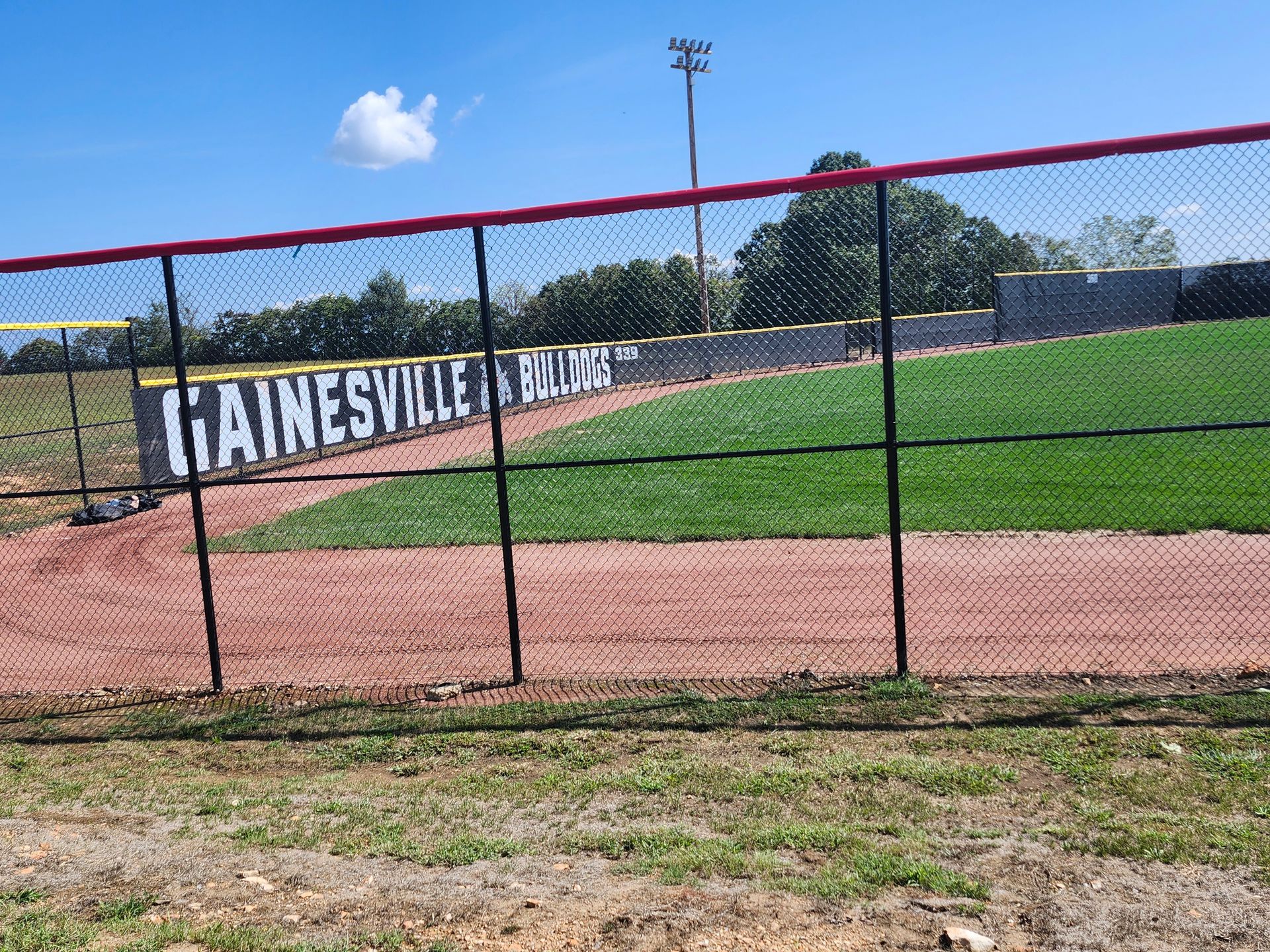 Baseball field behind a chain-link fence with a “GAINESVILLE” sign, green grass, and blue sky.
