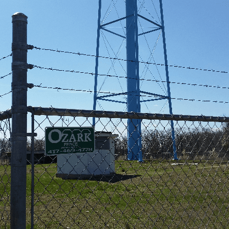A chain-link fence with barbed wire and an Ozark Fence sign by a metal tower.