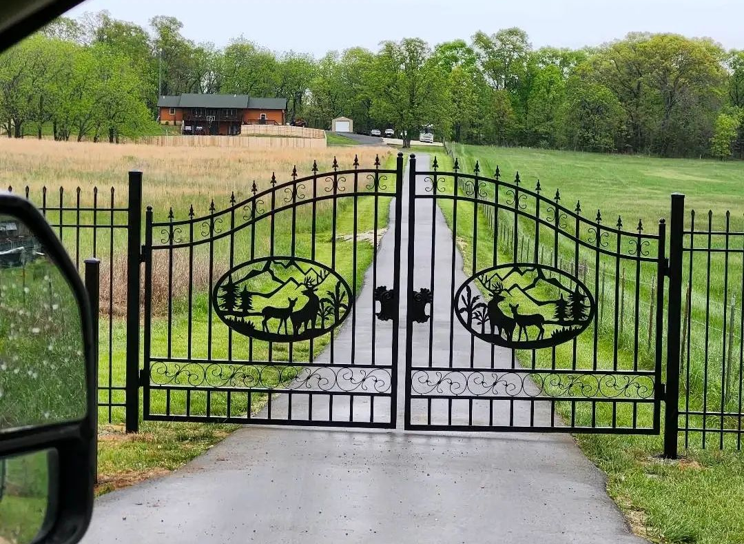 Black metal gate opening to a long gravel driveway across a grassy field with trees and a house in the distance