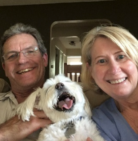 Man and woman smiling with white dog, all looking at the camera. Inside a home, the dog's tongue is out.
