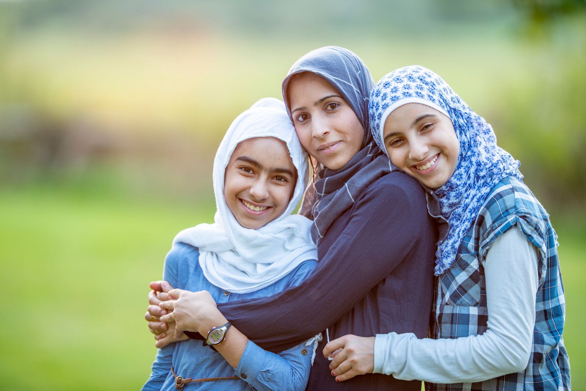 Three young women in hijab are hugging each other in a park.