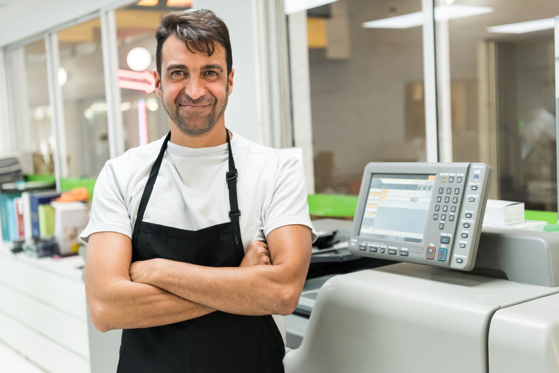 A man in an apron is standing in front of a printer with his arms crossed.