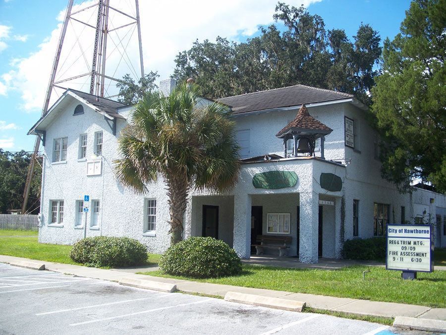 White stucco building with a bell tower and water tower in the background, surrounded by trees and grass.