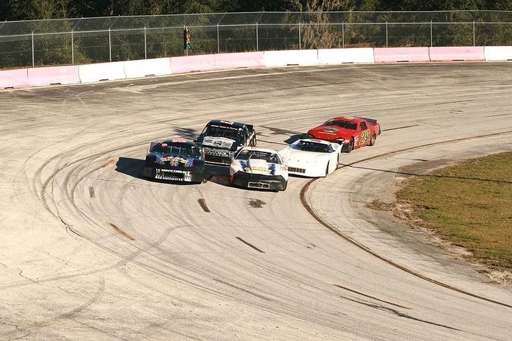 Race cars on a curved track, racing side-by-side. Tire marks on the track, fences in the background.