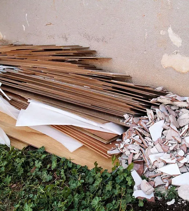 Pile of discarded cardboard, wood, and broken tiles next to a weathered wall and greenery.