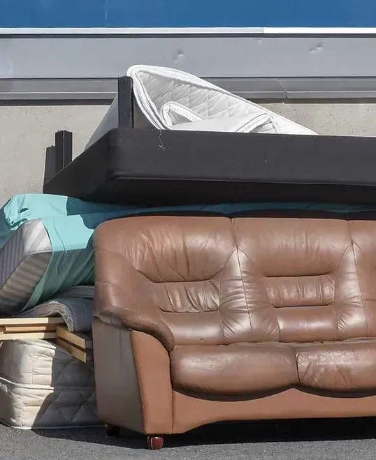 Pile of discarded furniture, including a brown leather sofa, mattresses, and a box spring, outdoors.