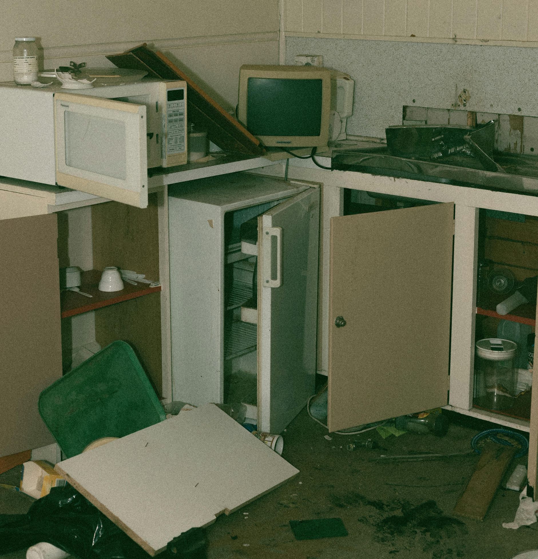 Messy, cluttered kitchen with open refrigerator, microwave, and cabinets. Debris and trash litter the floor.