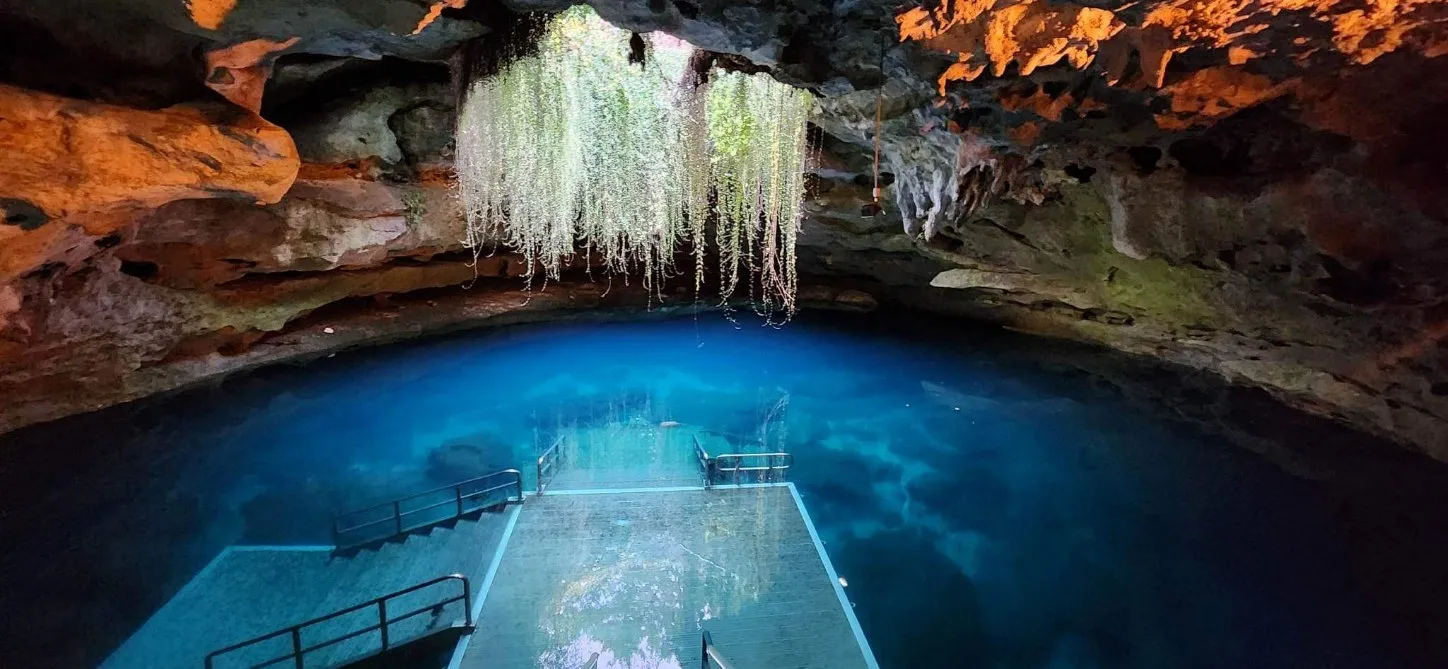 Blue water-filled cave with a dock and hanging white formations. Orange cave ceiling.