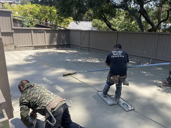 Two workers smoothing wet concrete on a large outdoor patio. Brown fence and trees in background.