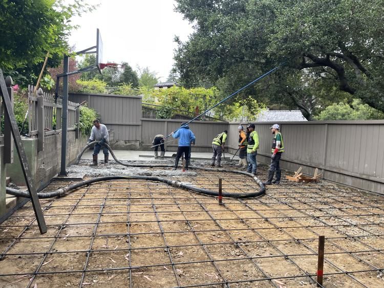 Construction workers pouring concrete for a patio. Rebar grid laid out, surrounded by a wooden fence, and trees.