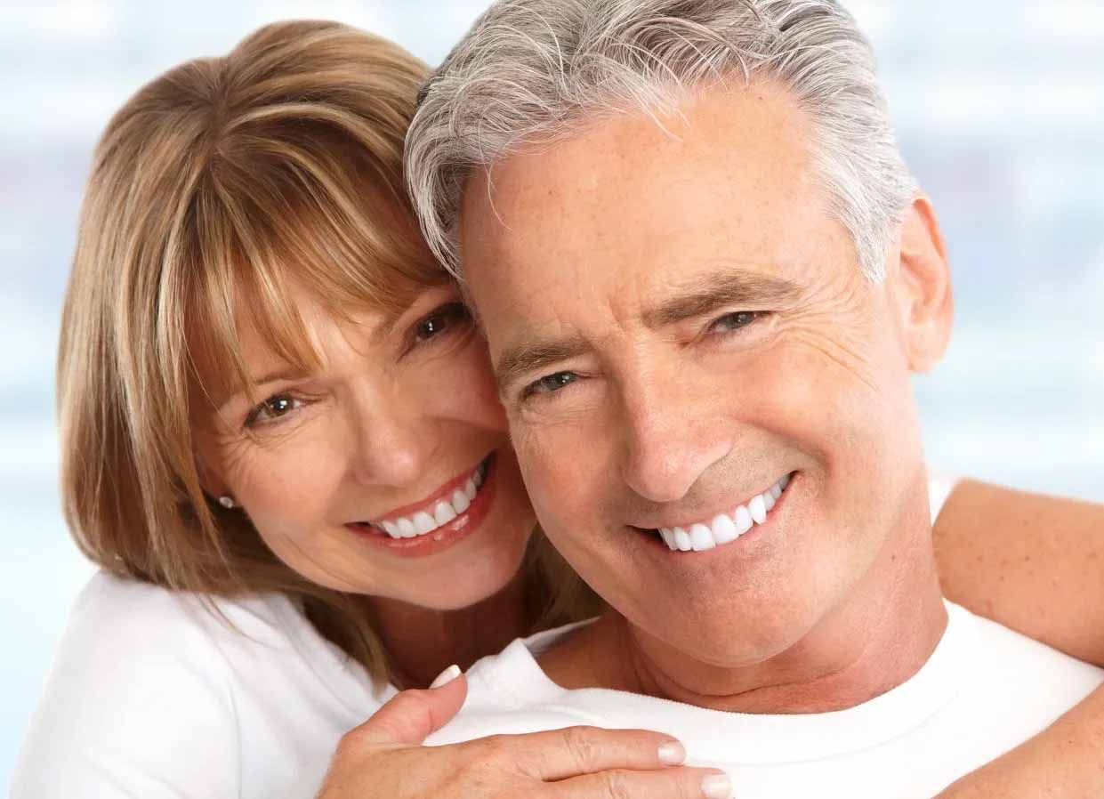 Couple souriant enlacé ; la femme entoure l’homme de ses bras. Tous deux portent des chemises blanches et affichent de larges sourires ; fond bleu clair.