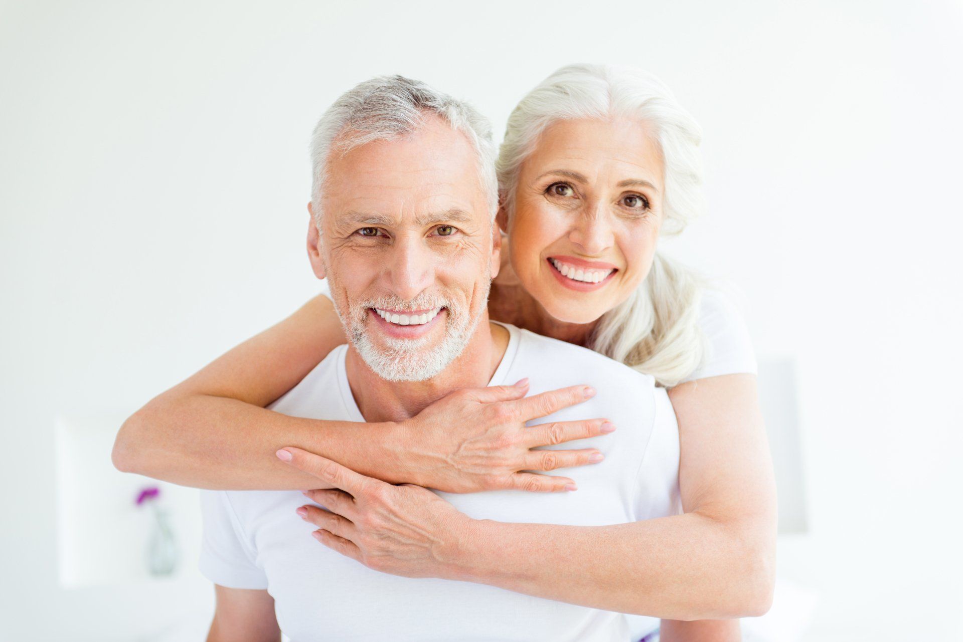 Un couple âgé souriant s'enlaçant, l'homme devant, la femme derrière, les bras autour de ses épaules.