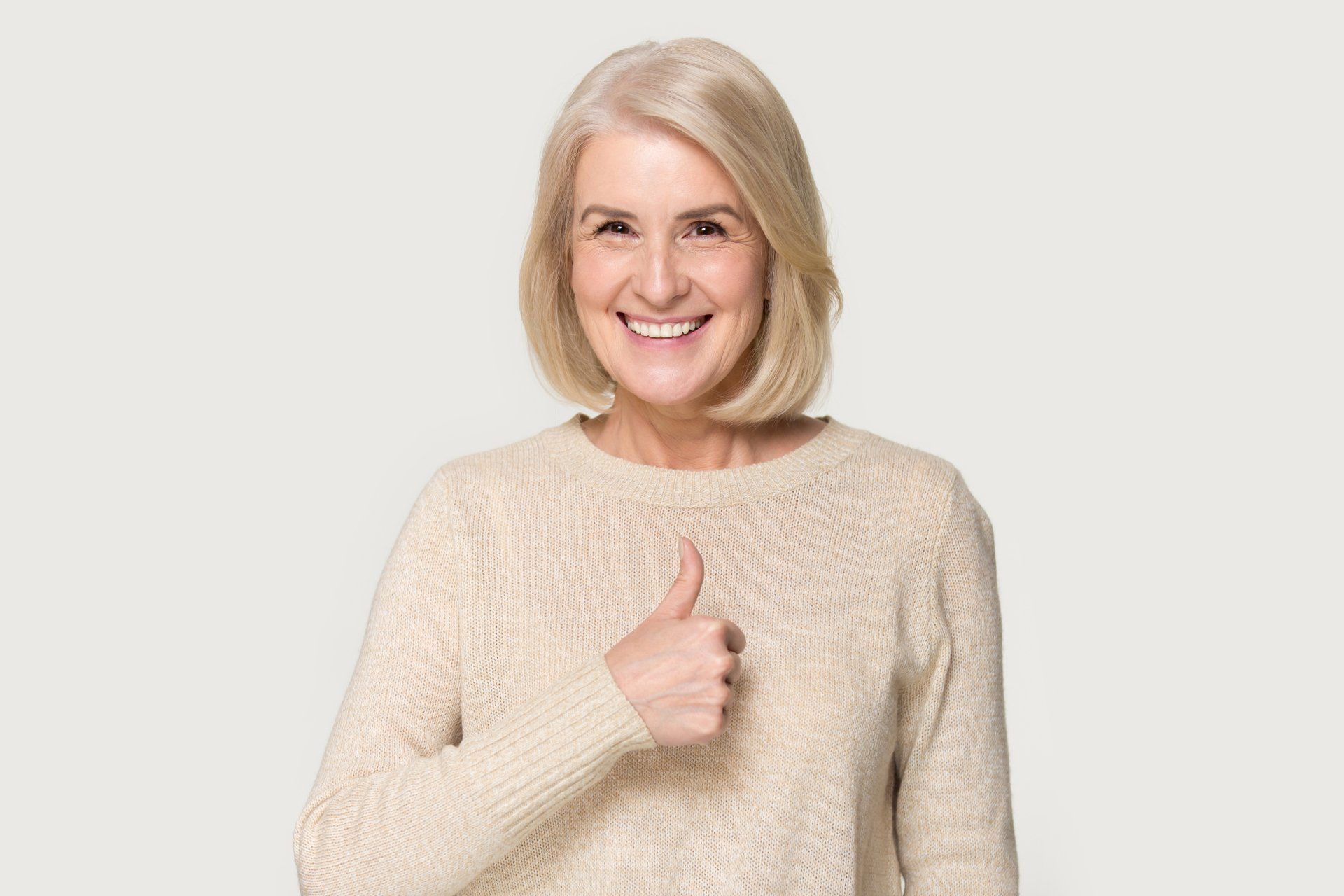 Femme aux cheveux blonds portant un pull crème, levant le pouce et souriant.