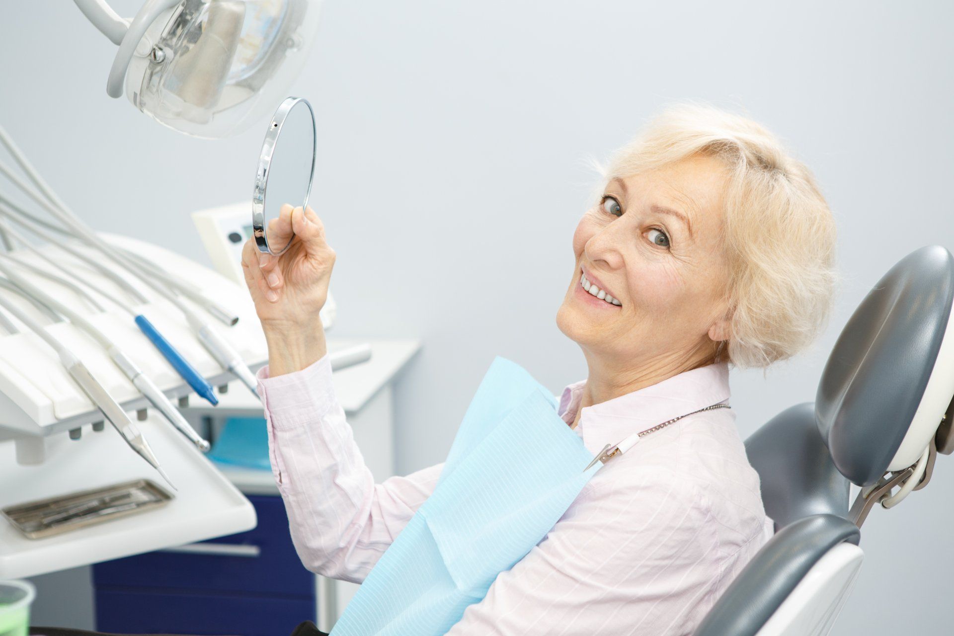 Une femme, assise sur un fauteuil de dentiste, sourit, un miroir à la main, en train d'examiner ses dents. Des instruments et du matériel dentaires sont visibles en arrière-plan.