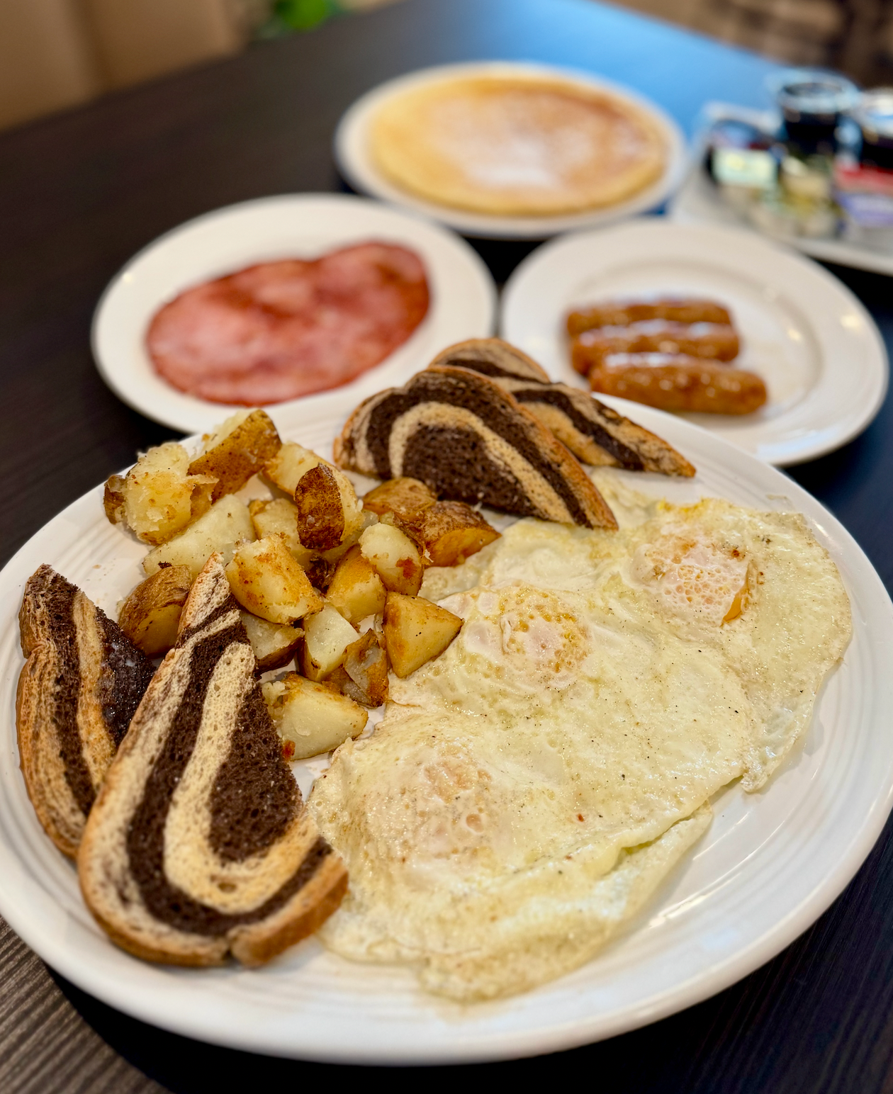 Breakfast plate with eggs, potatoes, toast, ham, sausage, and a cup of coffee in the background