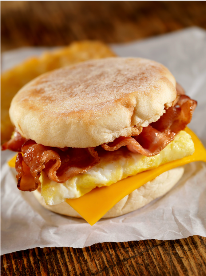 Breakfast sandwich with bacon, egg, and cheese on an English muffin, wrapped in white paper.