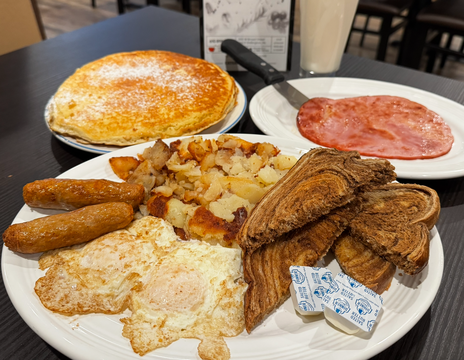 Breakfast spread with pancakes, eggs, bacon, sausage, potatoes, and toast on a table