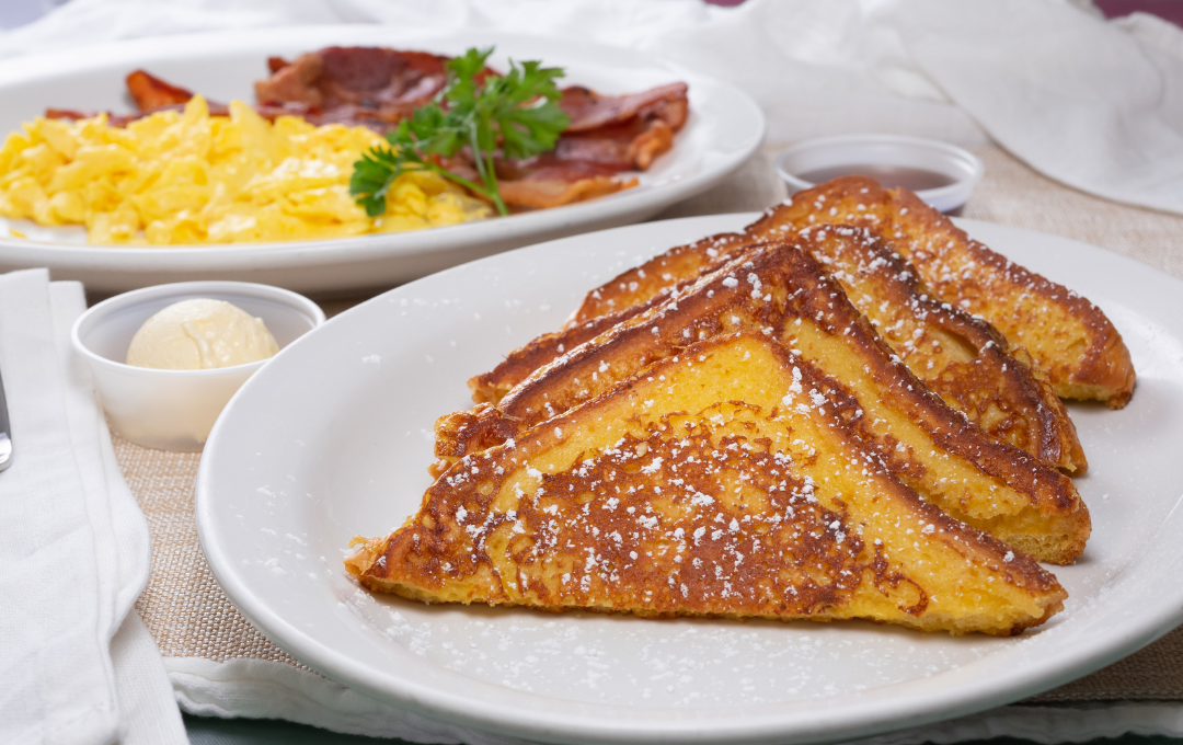 French toast topped with syrup on a white plate, with eggs and bacon in the background