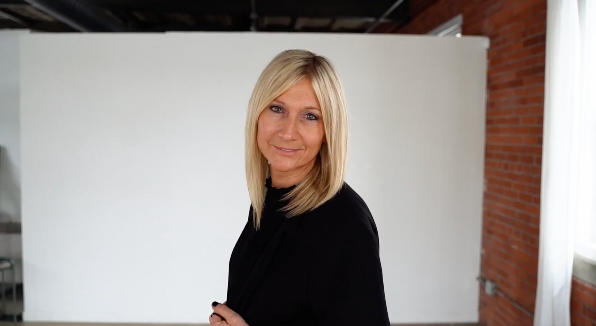 A woman in a black sweater is standing in front of a white wall.