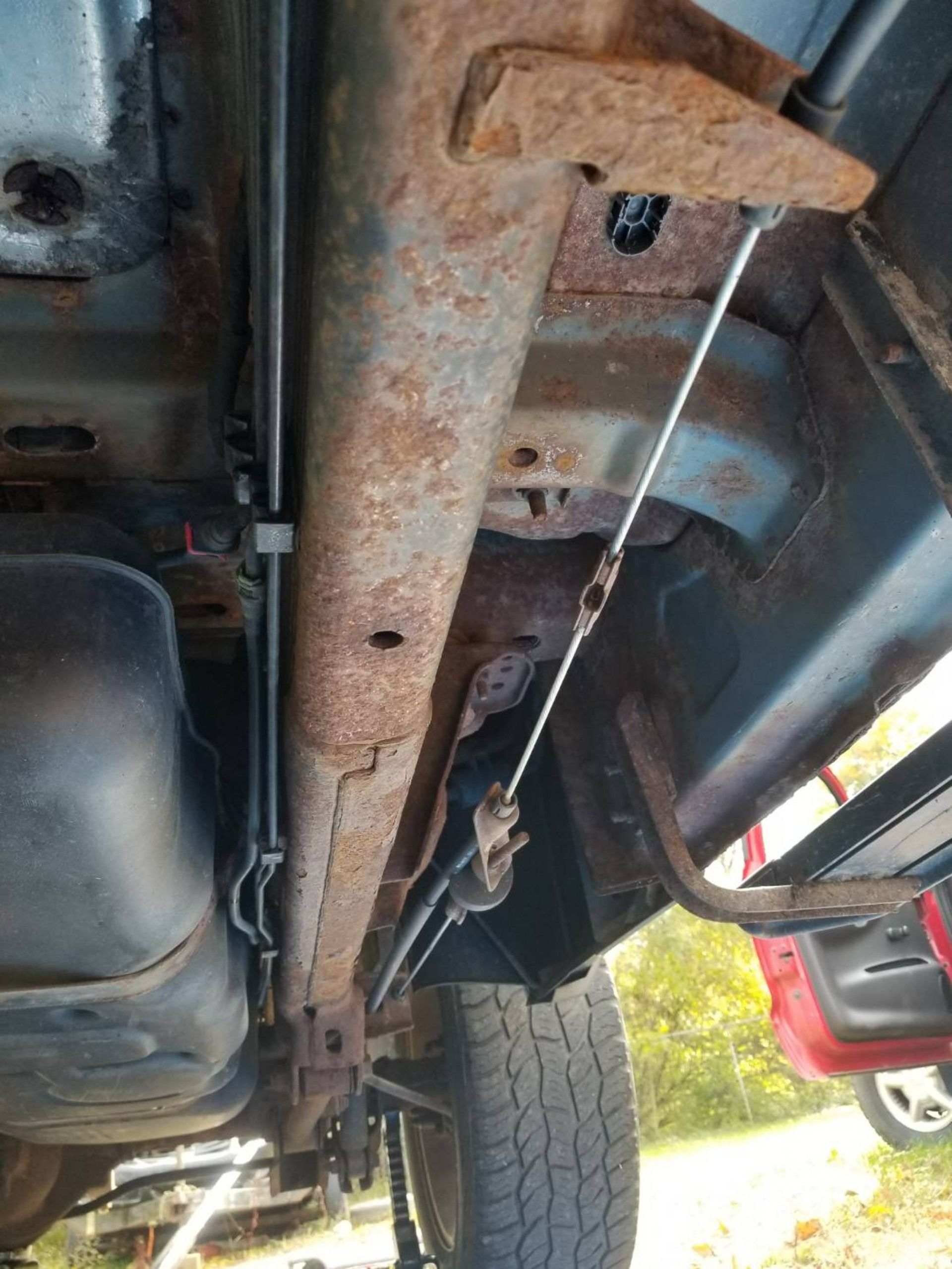 Undercarriage of a rusty vehicle showing frame, suspension parts, and wheel underneath