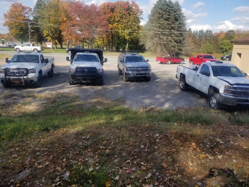 Five pickup trucks parked on a gravel lot beside trees with autumn leaves