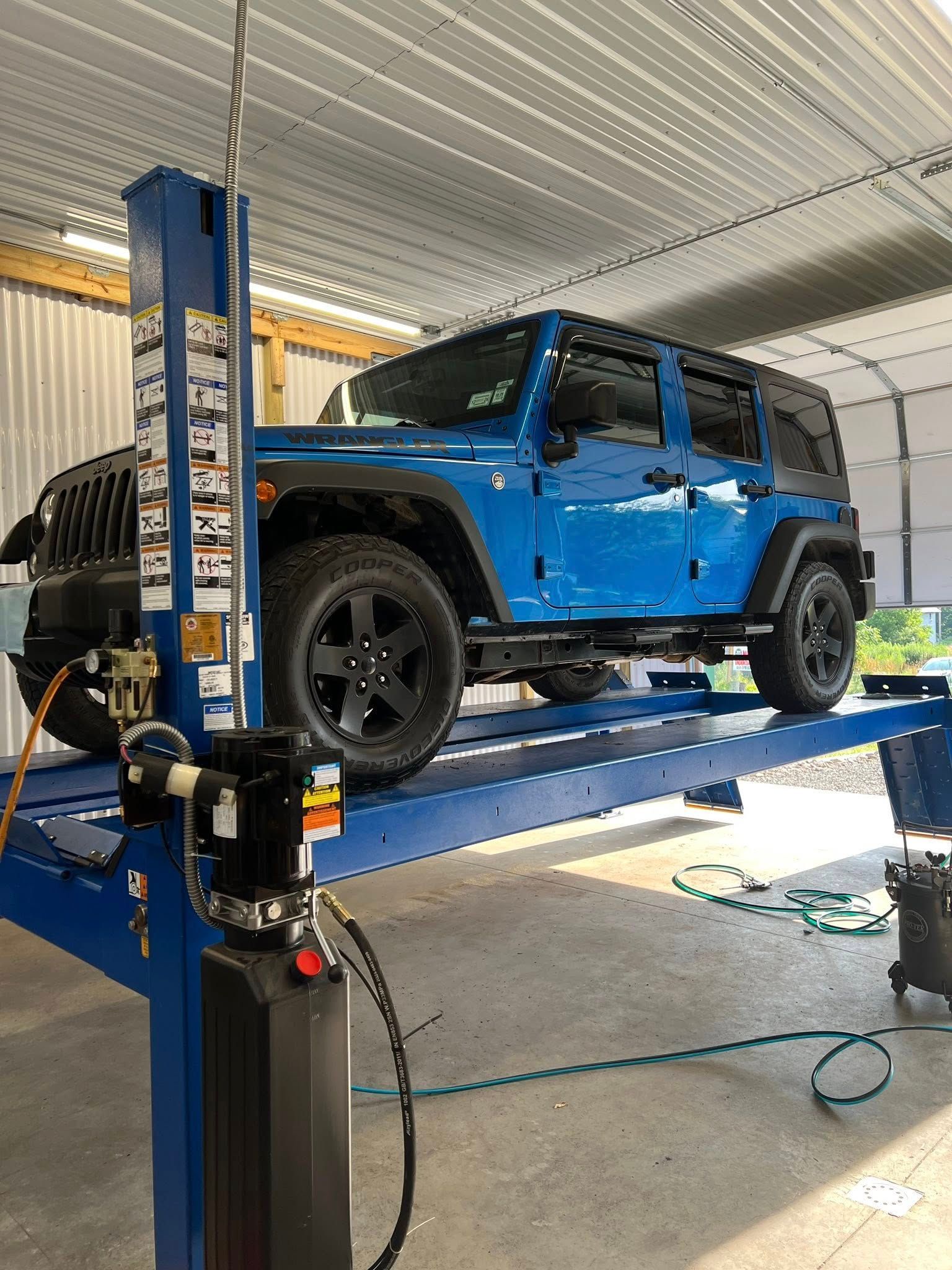 Blue Jeep Wrangler on a car lift in a garage with an open hood, viewed from the side.