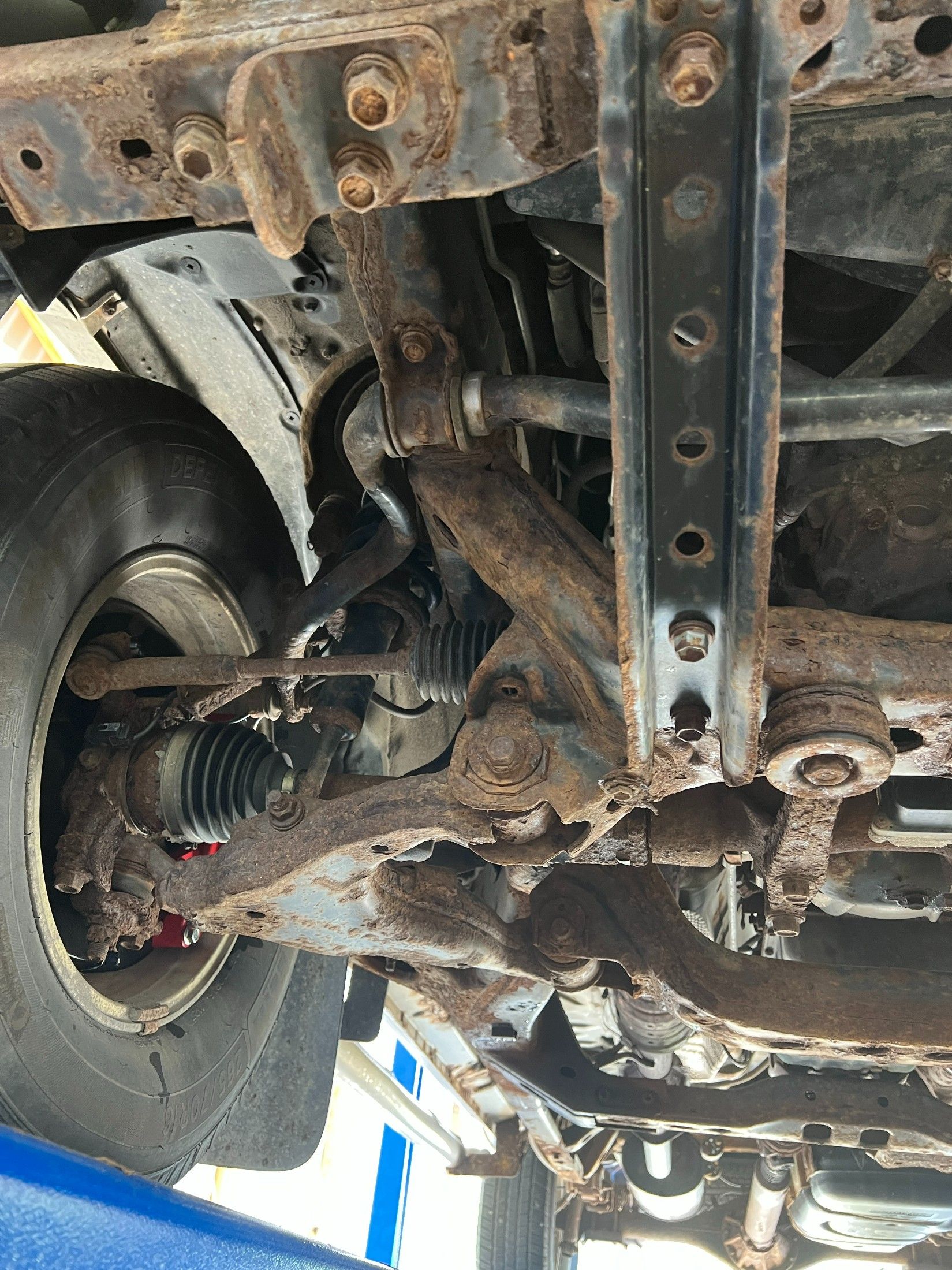 Undercarriage of a rusted vehicle, showing suspension, axle, and wheel assembly in a repair shop