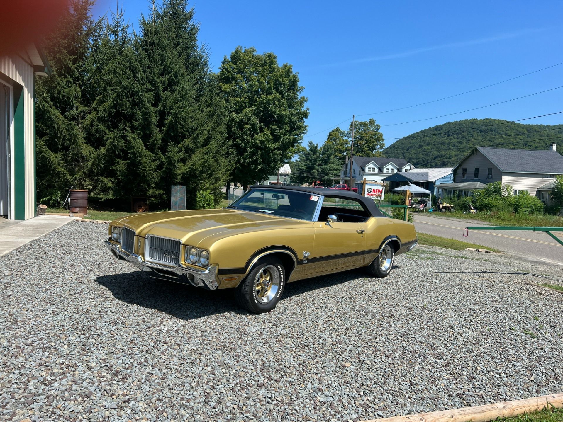 Gold classic convertible parked on gravel beside a building, with trees and hills in the background