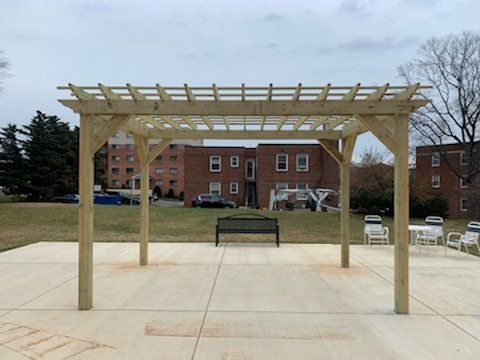 A wooden pergola with a bench underneath it in front of a brick building.