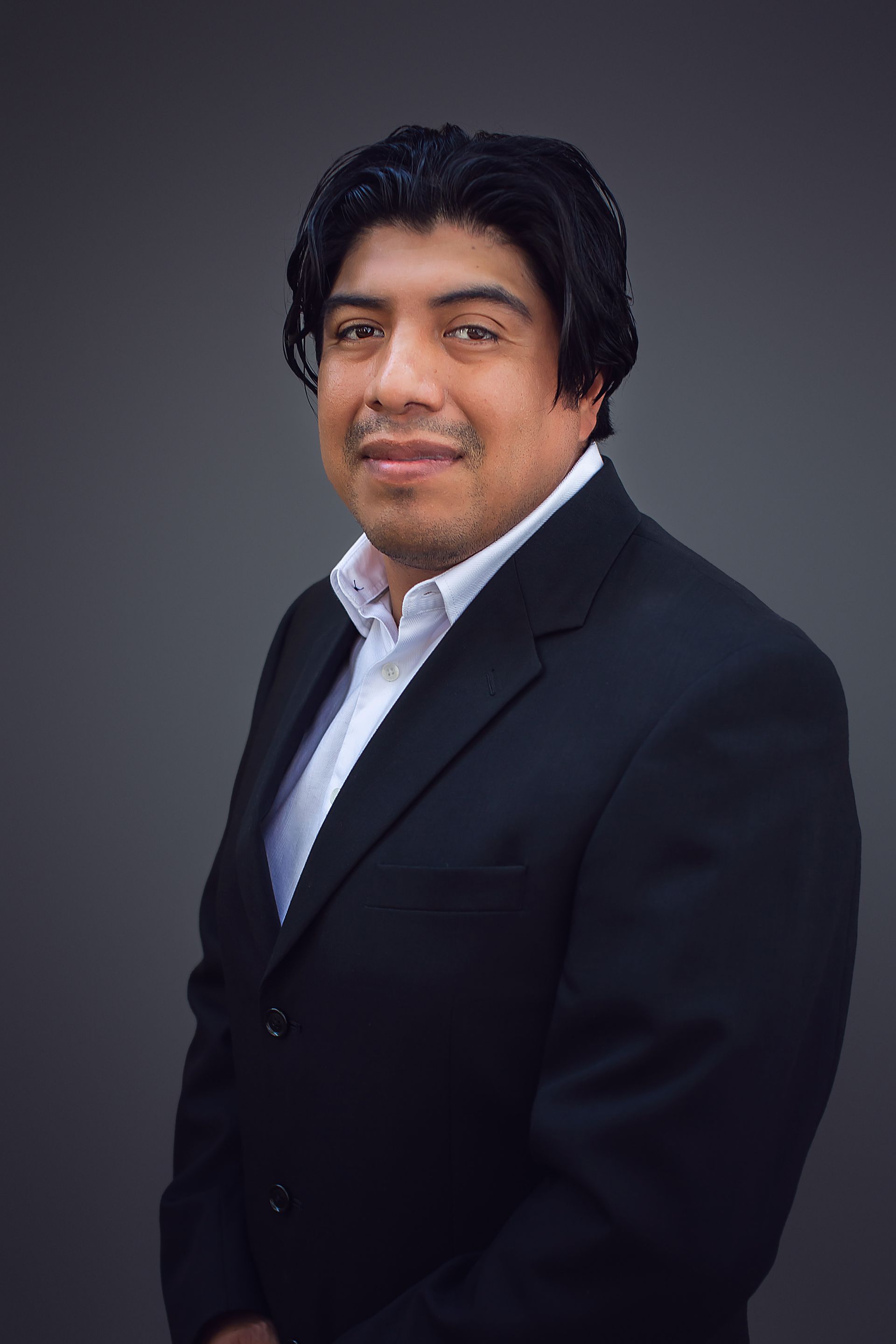 A man in a suit and white shirt is standing in front of a gray background.