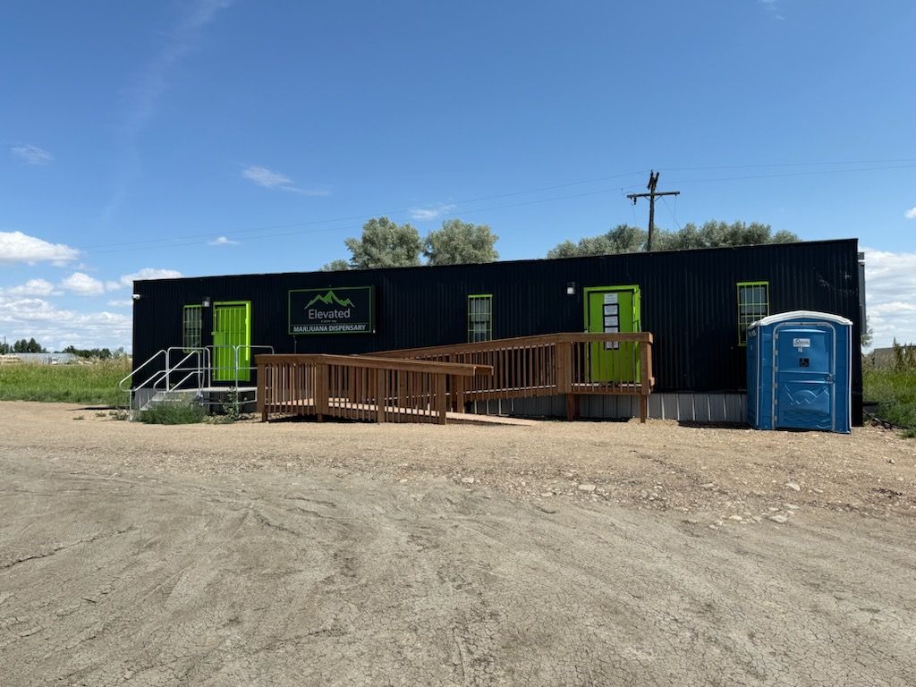 Black building with green doors, a wooden ramp, and a blue portable toilet on a gravel lot.