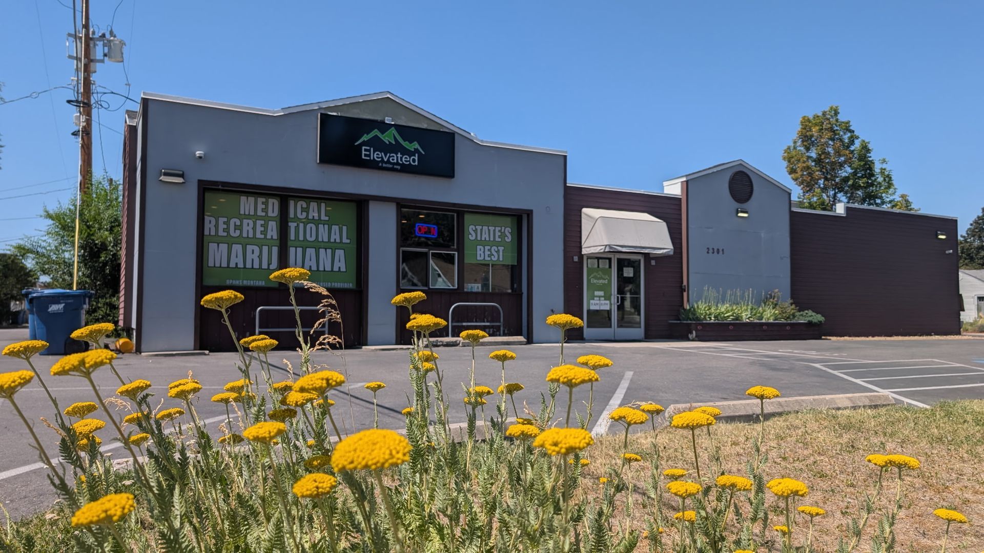 Exterior of a marijuana dispensary, gray building with a green logo, sunny day.