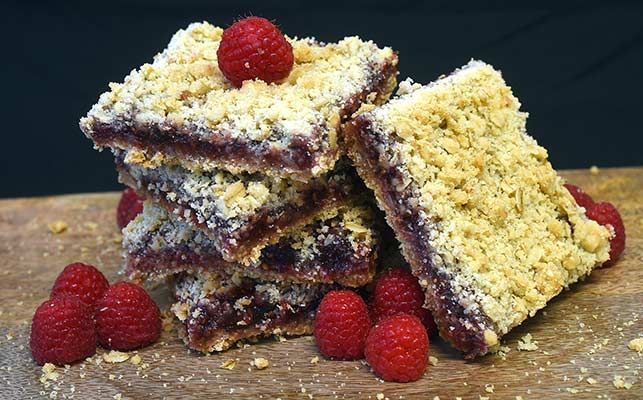 A stack of raspberry bars with raspberries on top on a wooden cutting board.