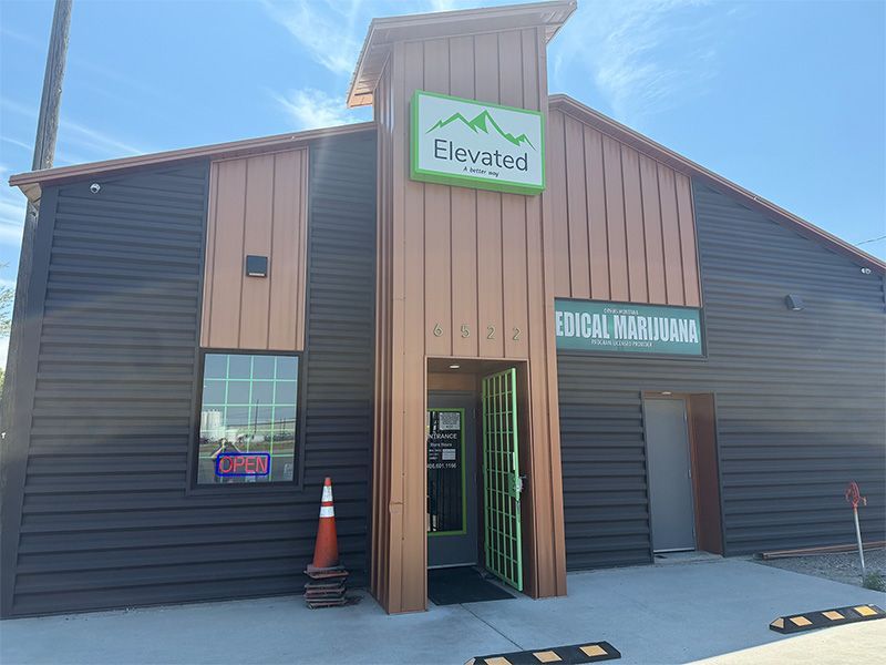 Exterior of Elevated dispensary with a green and white logo. Brown and black building with open door.