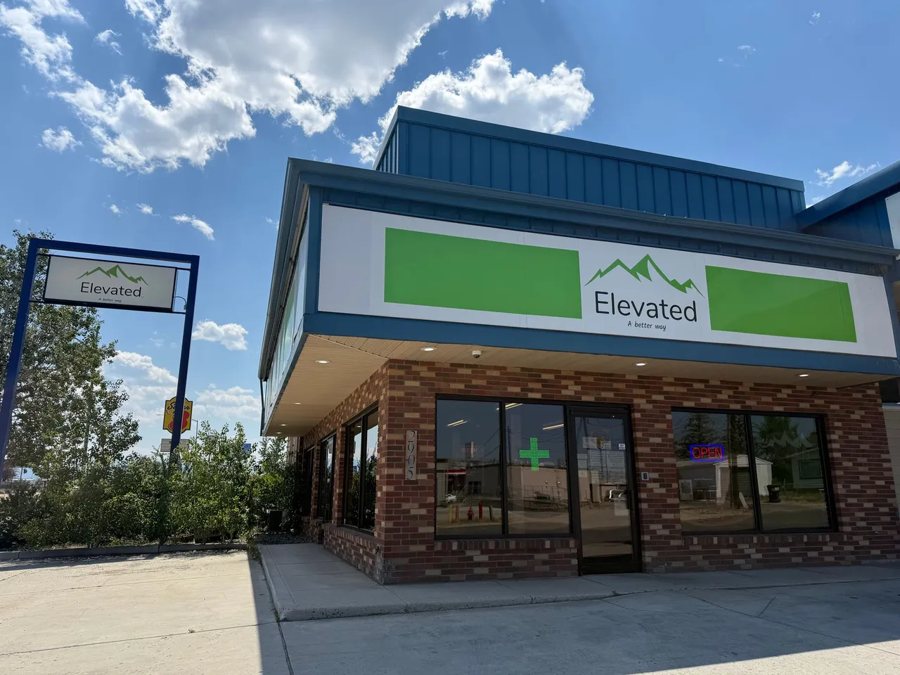 Elevated cannabis dispensary exterior. Brick building with green and blue accents, sign with logo, sunny day.