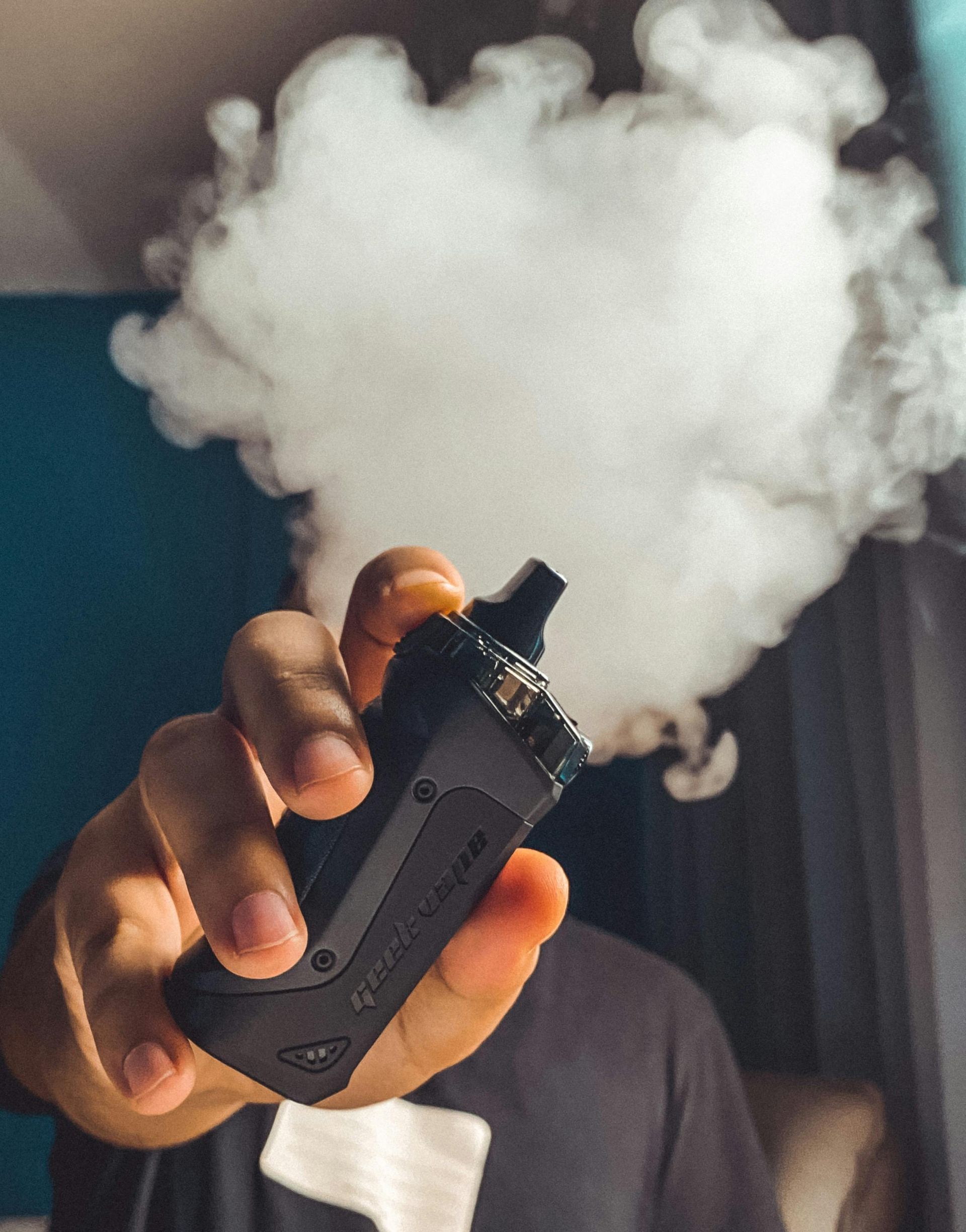 Person holding a black vape, exhaling a large cloud of white vapor.