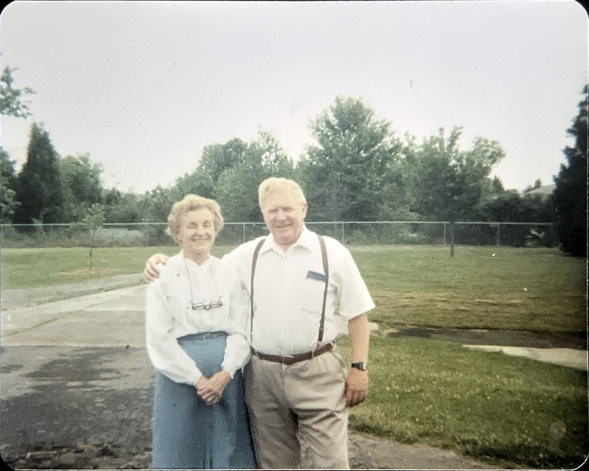 Two people standing together outdoors on a paved path near a grassy field and trees
